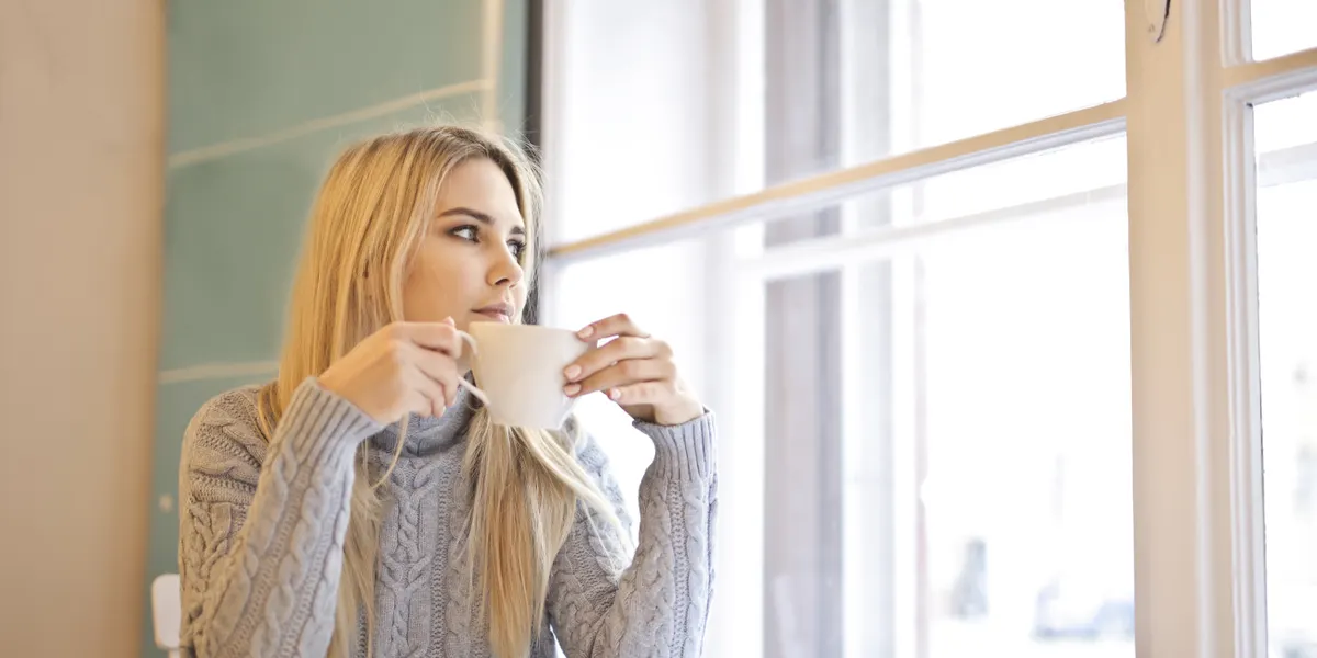 A woman in a grey knitted jumper holds a cup of coffee with both hands beside a bright window, looking out.
