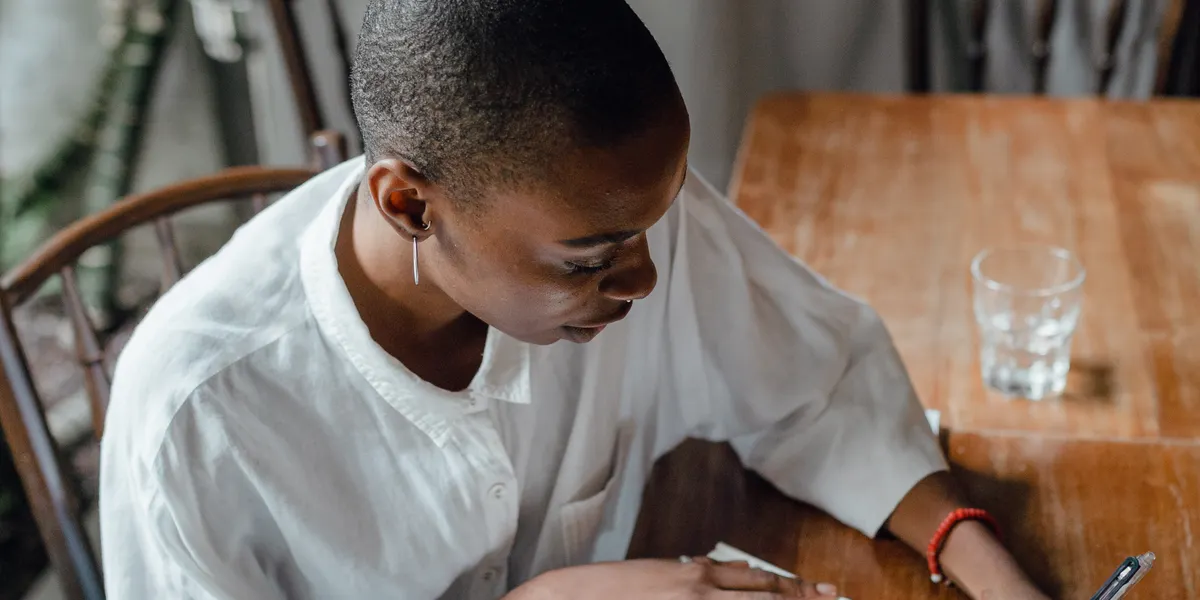 A woman in a white shirt sits at a sunlit wooden table, leaning over a notebook and writing.